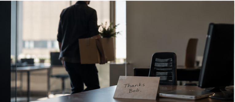 A man carrying a box out of an office, past a card on the desk that reads: Thanks Bob.